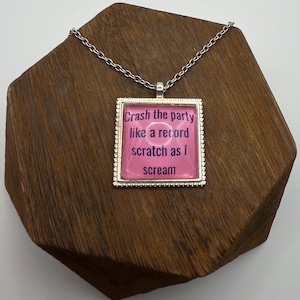 May include: A silver-toned necklace with a square pendant. The pendant has a pink background with the words "Crash the party like a record scratch as I scream" in purple text. The pendant is on a wooden surface.