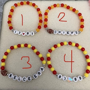Könnte beinhalten: Vier Armbänder aus roten und gelben Perlen, jedes mit einem anderen Design. Das erste Armband hat die Zahlen 13, ein Herz und 87. Das zweite Armband hat einen Fußball und das Wort "CHIEFS". Das dritte Armband hat einen Fußball und die Worte "KELCE ERA". Das vierte Armband hat die Zahlen 87, ein Herz und 89.