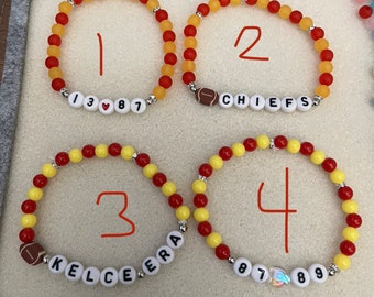 Op de afbeelding: Vier armbanden gemaakt met rode en gele kralen, elk met een ander ontwerp. De eerste armband heeft de nummers 13, een hartje en 87. De tweede armband heeft een voetbal en het woord "CHIEFS". De derde armband heeft een voetbal en de woorden "KELCE ERA". De vierde armband heeft de nummers 87, een hartje en 89.