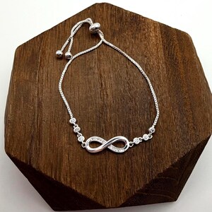 May include: A silver-toned bracelet featuring an infinity symbol adorned with small, clear gemstones. The adjustable chain bracelet is displayed on a dark brown, geometric wooden surface.