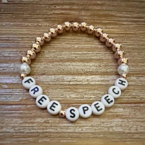 Könnte beinhalten: Ein Perlenarmband mit den Worten "FREE SPEECH" in weißen Buchstabenperlen. Das Armband hat goldfarbene Perlen und zwei perlenartige Perlen. Das Armband wird auf einer Holzoberfläche präsentiert.