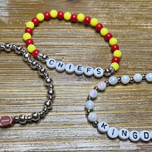 May include: Three beaded bracelets, one with red and yellow beads, one with silver beads and a football charm, and one with iridescent white beads. The bracelets spell out "CHIEFS" and "KINGDOM".