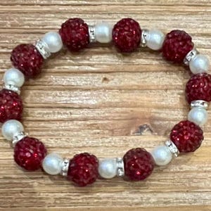 May include: A beaded bracelet featuring alternating red, sparkly beads and white, pearl-like beads. The red beads are clustered and surrounded by silver-colored spacers. The bracelet is displayed on a wooden surface.