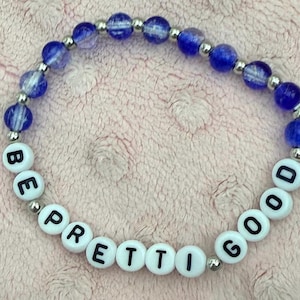 May include: A bracelet made of blue and silver beads. The bracelet features white letter beads spelling "BE PRETTI GOOD". The bracelet is displayed on a pink surface.