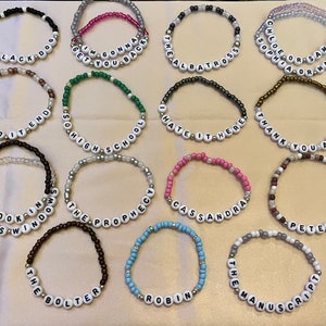 Puede incluir: Una colección de 15 pulseras de cuentas con varios colores y frases, incluyendo "The Black Dog", "Get You Back", "The Albatross", "How Did It End", "So High School", "I Hate It Here", "Thank You Aimee", "Peoples Windows", "The Prophecy", "Cassandra", "Peter", "The Bolter", "Robin", y "The Manuscript".