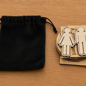May include: A black drawstring pouch beside a stack of wooden gender symbol cutouts. The cutouts are in the shape of a stick figure woman and man. The background is a light brown wooden surface.