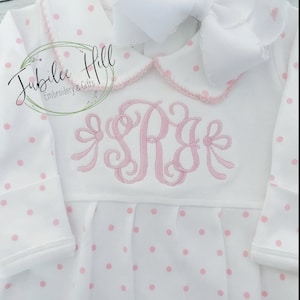 May include: White baby gown with pink polka dots and a pink embroidered monogram. The gown has a white Peter Pan collar with a pink trim.
