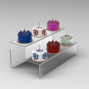Könnte beinhalten: Ein zweistufiger Acryl-Kuchenständer mit fünf dekorierten Kuchen. Die Kuchen sind in verschiedenen Farben und Designs dekoriert, darunter Blau, Rot, Weiß und Grün. Jeder Kuchen hat eine einzelne rosa Kerze.