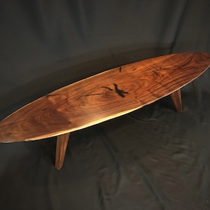 Puede incluir: Una mesa de centro de madera ovalada y larga con veta de madera marrón oscuro y tres patas. La mesa está hecha de madera de nogal y tiene un acabado natural.