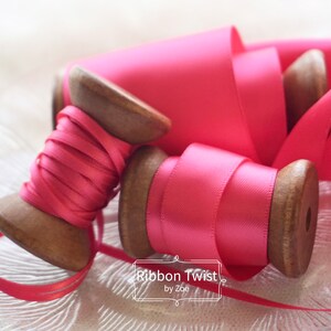 May include: Two spools of pink satin ribbon on a white surface. The spools are made of wood and have a pink ribbon wound around them. The text "Ribbon Twist by Zoe" is visible in the lower right corner of the image.