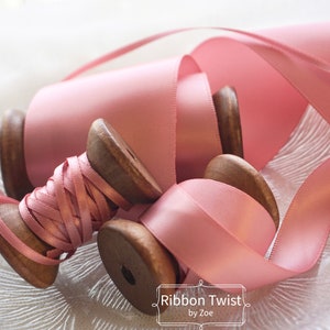 May include: Two spools of pink satin ribbon, one on a wooden spool and the other on a larger wooden spool. The ribbon is wound around the spools and the ends are loose. The spools are sitting on a white, textured surface. The text "Ribbon Twist by Zoe" is visible at the bottom of the image.