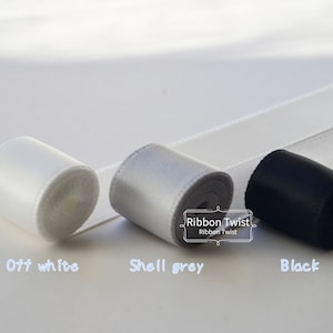 May include: Three spools of ribbon in off-white, shell grey, and black. The ribbon is rolled up and the ends are visible. The text "Ribbon Twist" is printed on a white label.