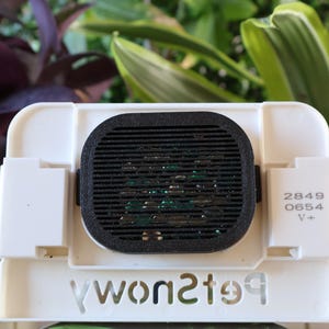 May include: A white rectangular device with a black, oval-shaped vent. The device has the text "petowsky" in a cut-out design. The numbers "2849 0654 V+" are printed on the side. Green and purple plants are in the background.