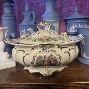 May include: A white porcelain serving dish with a lid, adorned with gold trim and floral accents. The dish features a detailed painting of figures and is surrounded by blue ceramic vases and jars.