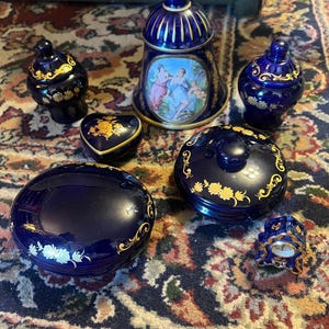 Op de afbeelding: Een verzameling kobaltblauwe porseleinen decoratieve items met gouden bloemaccenten. Inclusief een bel, kleine potjes met deksel en een hartvormige sieradendoos. De bel heeft een geschilderde scène van figuren.