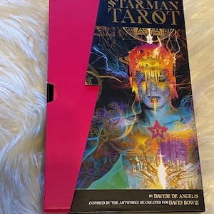 以下が含まれることがあります： 鮮やかなイラストが特徴のタロットカードボックスセット。ピンクと黒の組み合わせで、「STARMAN TAROT」のタイトルが前面に表示されています。デヴィッド・ボウイにインスパイアされたアートワークです。