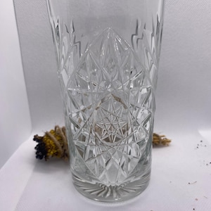 May include: A clear, patterned drinking glass with a diamond-shaped design. The glass has a cylindrical shape and a detailed, textured pattern. The glass is empty and sits on a white surface.