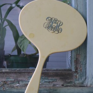May include: A vintage, oval-shaped hand mirror in a pale yellow hue. The mirror features a long handle and an ornate monogram in blue script. The mirror is resting on a weathered wooden surface.