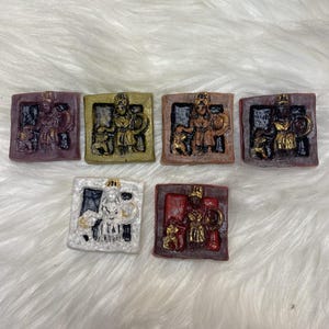 Puede incluir: Colección de seis pequeñas esculturas en relieve cuadradas. Cada una presenta una figura clásica en un marco cuadrado. Las esculturas son de varios colores, incluyendo burdeos, dorado y bronce, con un fondo blanco.