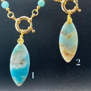 Op de afbeelding: Twee goudkleurige kettingen met edelsteenhangers. Elke ketting heeft een ovale hanger met blauwe, witte en bruine tinten. De hangers hangen aan een gouden ketting met een grote sluiting. Eén ketting heeft kleine blauwe kralen.