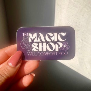 K&ouml;nnte beinhalten: Ein lilafarbener Aufkleber mit wei&szlig;em Text, der "THE MAGIC SHOP WILL COMFORT YOU" lautet. Der Text ist in einer stilisierten Schriftart gehalten und von zwei wei&szlig;en Blumen umgeben.