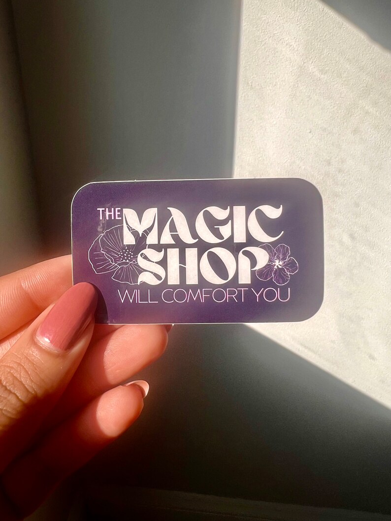 K&ouml;nnte beinhalten: Ein lilafarbener Aufkleber mit wei&szlig;em Text, der "THE MAGIC SHOP WILL COMFORT YOU" lautet. Der Text ist in einer stilisierten Schriftart gehalten und von zwei wei&szlig;en Blumen umgeben.