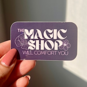 K&ouml;nnte beinhalten: Ein lilafarbener Aufkleber mit wei&szlig;em Text, der "The Magic Shop Will Comfort You" lautet. Der Text ist in einer Retro-Schriftart gehalten und von zwei wei&szlig;en Blumen umgeben.