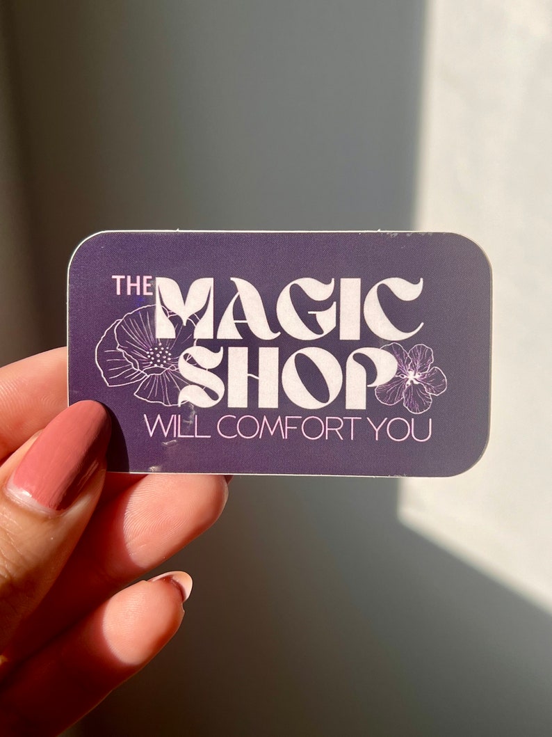 K&ouml;nnte beinhalten: Ein lilafarbener Aufkleber mit wei&szlig;em Text, der "The Magic Shop Will Comfort You" lautet. Der Text ist in einer Retro-Schriftart gehalten und von zwei wei&szlig;en Blumen umgeben.
