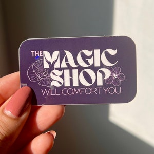 K&ouml;nnte beinhalten: Ein lilafarbener Aufkleber mit wei&szlig;em Text, der "The Magic Shop Will Comfort You" lautet. Der Text ist in einer Retro-Schriftart gehalten und von zwei wei&szlig;en Blumen umgeben.