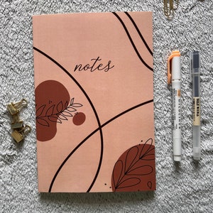 May include: A pink notebook with the word "notes" written on the front cover. The cover has a brown and black abstract design with leaves and curved lines.