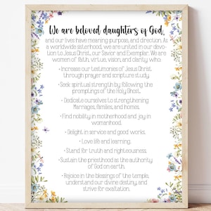 May include: A framed print with the text "We are beloved daughters of God." The text is surrounded by a floral border of blue, yellow, and purple flowers. The print is displayed on a white wall.