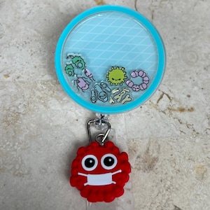 May include: A retractable badge reel with a clear, round top and a light blue background with a diamond pattern. The reel contains cartoon germs. A red, germ-shaped charm with a white face mask is attached.