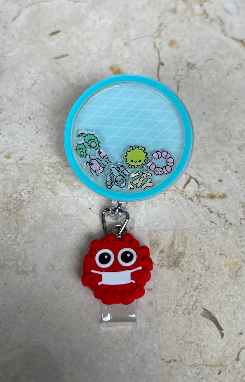 May include: A retractable badge reel with a clear, round top and a light blue background with a diamond pattern. The reel contains cartoon germs. A red, germ-shaped charm with a white face mask is attached.