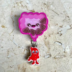 May include: A pink and light purple badge reel featuring a cartoon illustration of intestines with smiling faces and brown poop emojis. A red, sad-faced stomach charm hangs below.