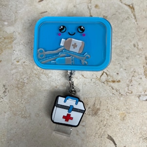 May include: A blue badge reel with a cartoon design featuring medical tools and a smiling face. The reel has a small white and red first aid kit charm attached. The design includes a syringe, scissors, and a bandage.