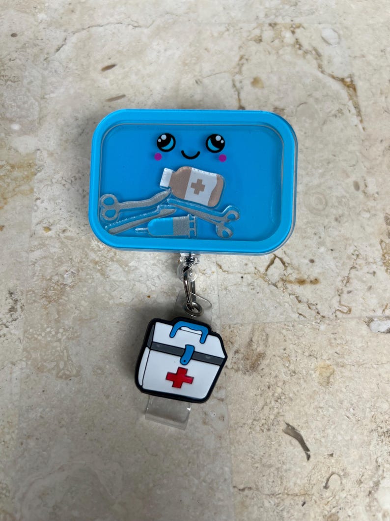 May include: A blue badge reel with a cartoon design featuring medical tools and a smiling face. The reel has a small white and red first aid kit charm attached. The design includes a syringe, scissors, and a bandage.