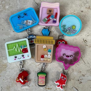May include: A collection of novelty badge reels with various designs. The reels include a blue surgical tray, a pink container with a biohazard symbol, a clear reel with blue and green elements, a monitor with a heartbeat, a pill bottle, and other medical-themed designs.