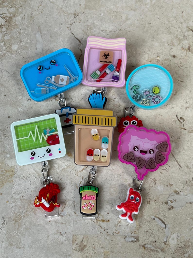 May include: A collection of novelty badge reels with various designs. The reels include a blue surgical tray, a pink container with a biohazard symbol, a clear reel with blue and green elements, a monitor with a heartbeat, a pill bottle, and other medical-themed designs.