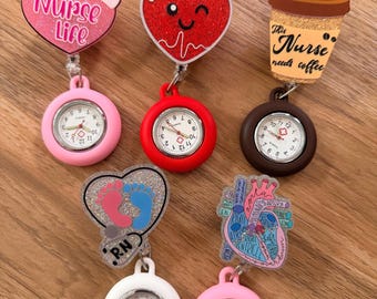 Puede incluir: Una colección de siete carretes de insignia con temática de enfermería con esferas de reloj. Los adornos incluyen un corazón rosa con "Nurse Life", un corazón rojo con una cara sonriente, una taza de café con "This Nurse needs coffee".