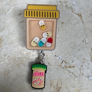 May include: A badge reel featuring a tan pill bottle design with a yellow cap and a clear window filled with colorful, smiling pill-shaped charms. A smaller charm with the word "chill" and pills hangs below.