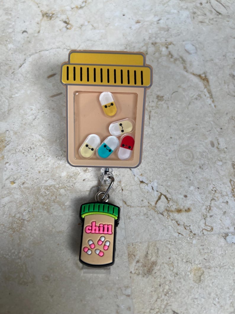 May include: A badge reel featuring a tan pill bottle design with a yellow cap and a clear window filled with colorful, smiling pill-shaped charms. A smaller charm with the word "chill" and pills hangs below.