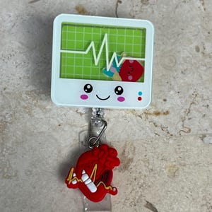 May include: A white badge reel with a green screen displaying a heartbeat monitor graphic. A red heart charm with a white line and a heartbeat graphic hangs below. The badge reel has a smiling face.