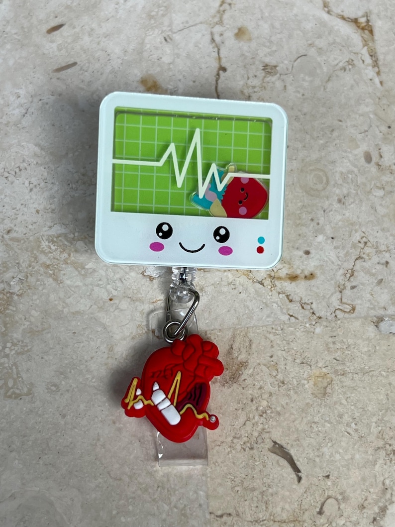 May include: A white badge reel with a green screen displaying a heartbeat monitor graphic. A red heart charm with a white line and a heartbeat graphic hangs below. The badge reel has a smiling face.