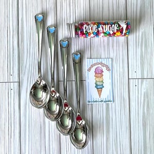 May include: Four silver-toned metal spoons with small blue accents on the handles, a clear plastic bottle filled with colorful star-shaped sprinkles labeled Pixie Sugar, and a rectangular card featuring an ice cream cone illustration.
