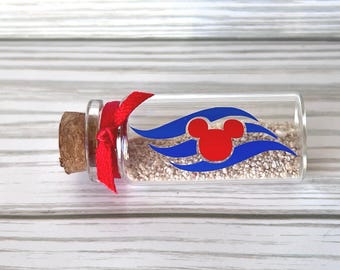 May include: A small clear glass bottle filled with light-coloured sand, sealed with a cork stopper and tied with a red ribbon. The exterior of the glass features a graphic design of a red circular icon with two smaller circles on top, centred between two blue wavy lines.