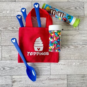 May include: A red tote bag with the word "Toppings" and a white ice cream graphic. The bag contains blue spoons with a Mickey Mouse head design. A clear jar of star-shaped sprinkles and a tube of M&M's are also present.
