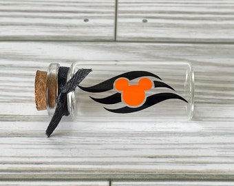 May include: A small, clear glass bottle with a cork stopper and a black ribbon tied around the neck. The bottle features an orange Mickey Mouse head logo with black wave accents. The bottle is lying horizontally.