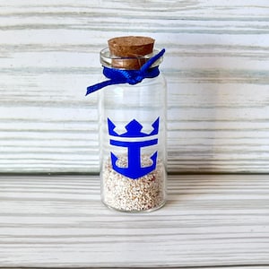 May include: A clear glass bottle with a cork stopper and a blue ribbon tied around the neck. The bottle features a blue anchor logo and is filled with small, light-colored granules. The background is a light wood.