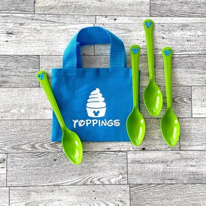 May include: A blue tote bag with a white ice cream design and the word "TOPPINGS" in white. Four green spoons with a blue Mickey Mouse head design are arranged around the bag. The bag has a blue handle.