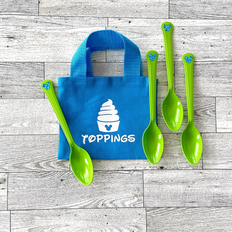 May include: A blue tote bag with a white ice cream design and the word "TOPPINGS" in white. Four green spoons with a blue Mickey Mouse head design are arranged around the bag. The bag has a blue handle.